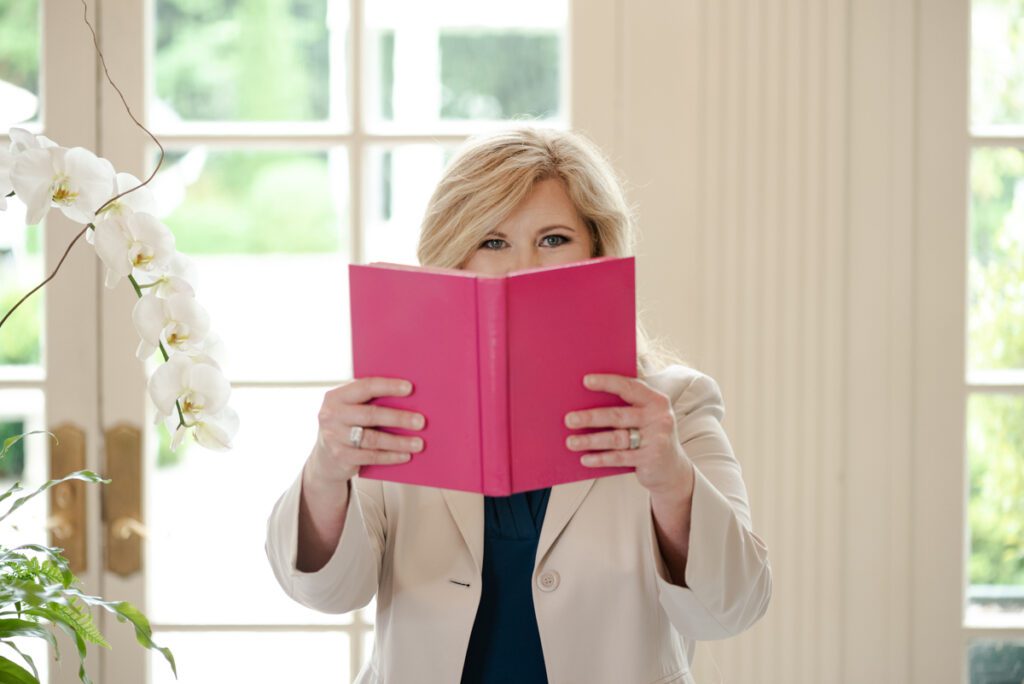 Woman holds a pink book in front of her face only showing her eyes.
