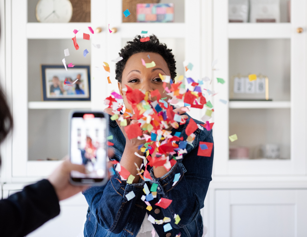 A hand holds a phone while taking a picture of a woman blowing confetti.